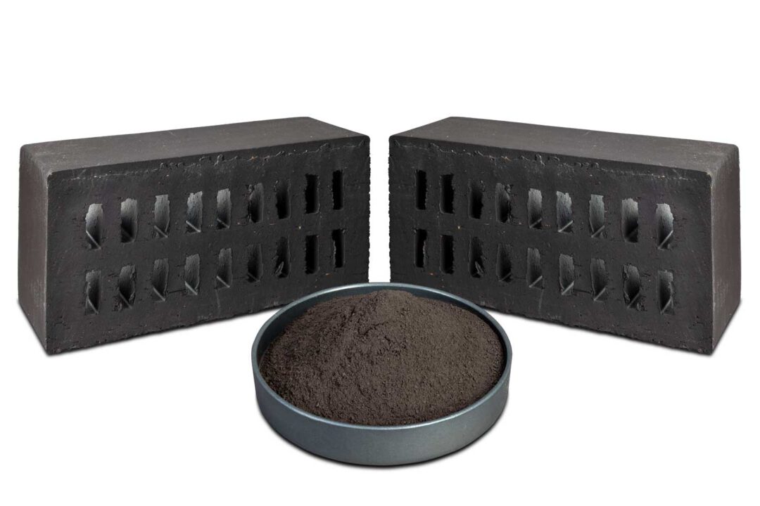 Schwarze Mahlschamotte aus Ziegelsteinen in einer dunklen Schale, dahinter zwei schwarze Ziegelsteine.